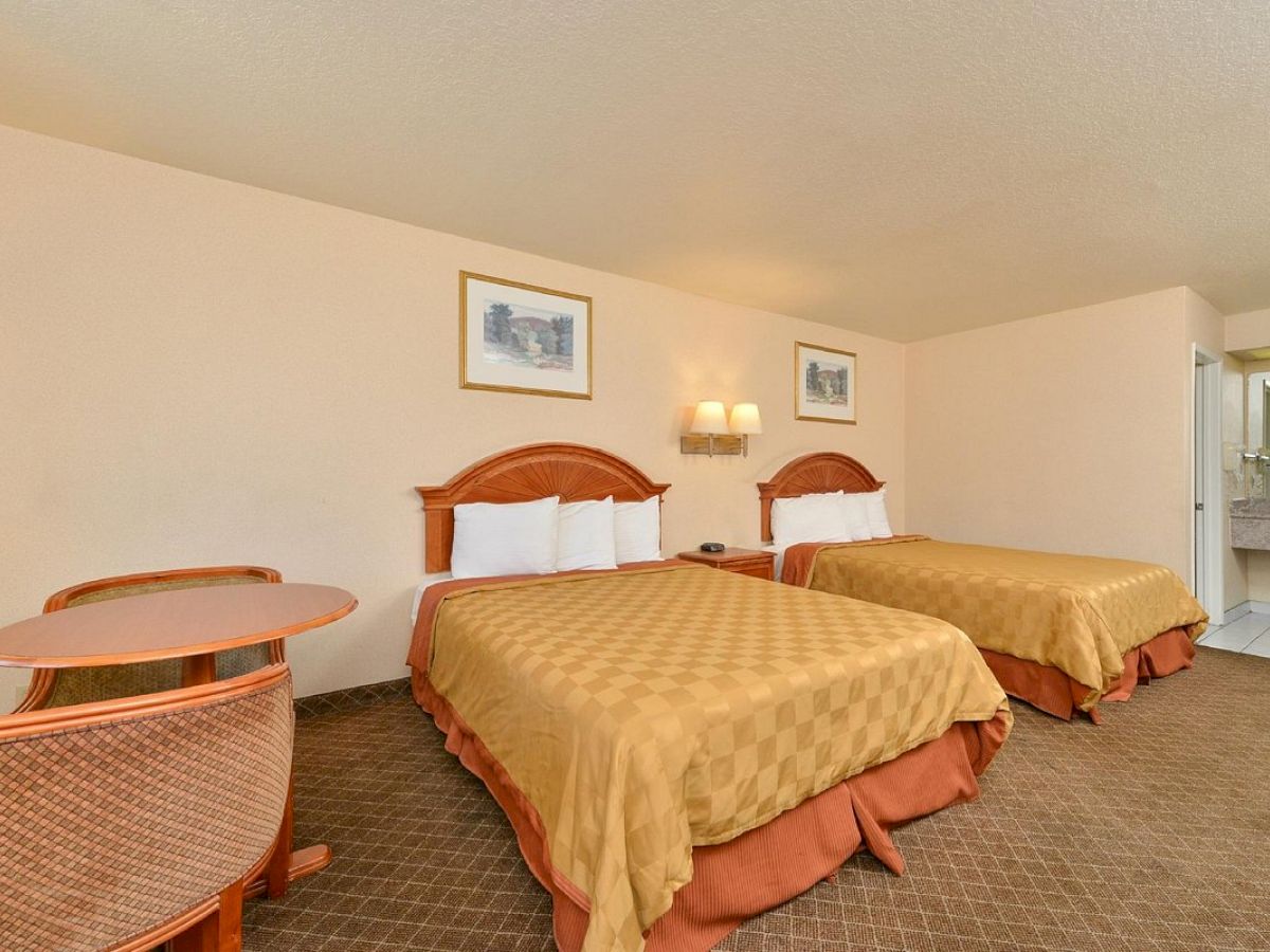 The image shows a hotel room with two double beds, a small round table with chairs, framed pictures on the wall, and a visible bathroom area.