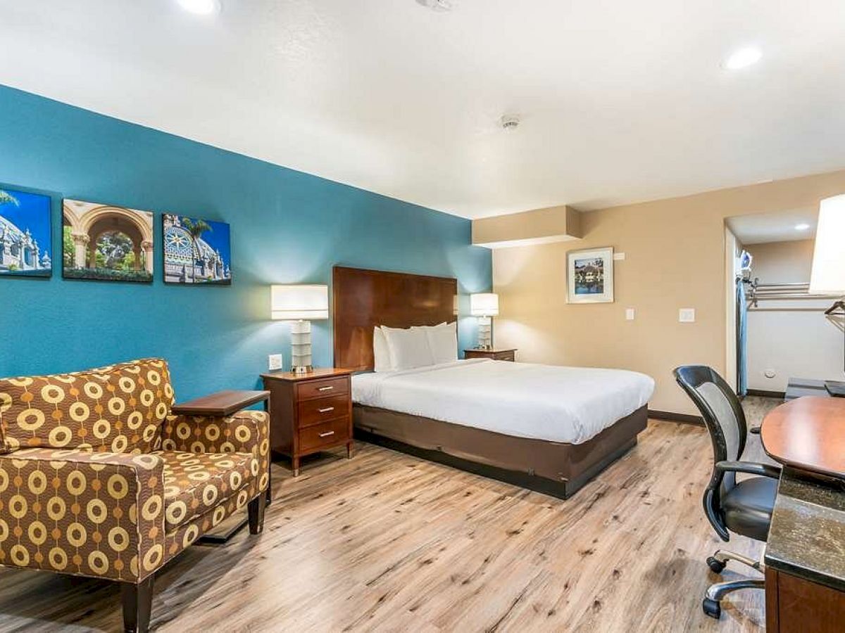 A modern hotel room featuring a double bed, a patterned armchair, a desk with a chair, and framed artwork on the walls, fully described.