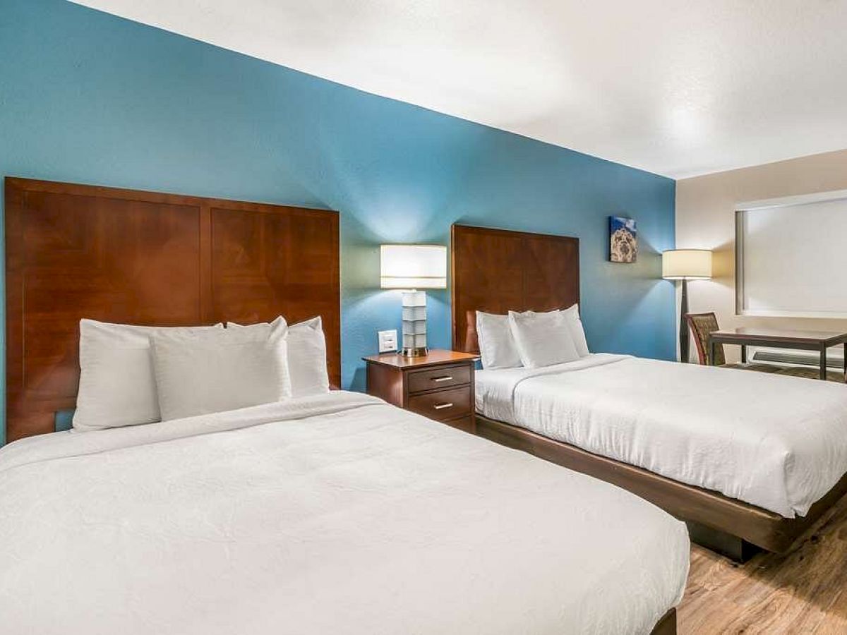 A hotel room with two double beds, a nightstand with a lamp, blue accent wall, and a small table and chairs in the corner, all well-lit.