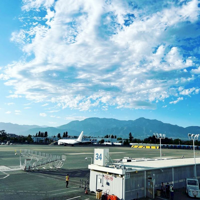 An airport with a runway and terminal, an airplane, vehicles, and mountains in the background under a partly cloudy sky.