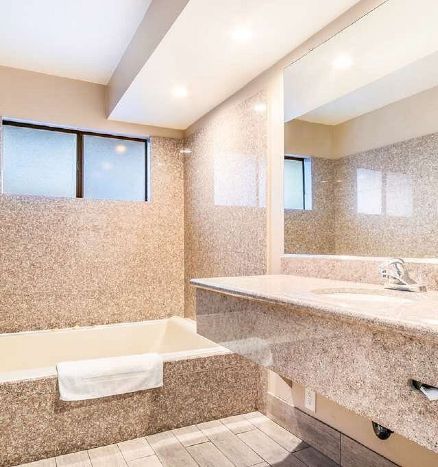 This image shows a modern bathroom with a bathtub, large mirror, granite countertops, and two frosted windows.