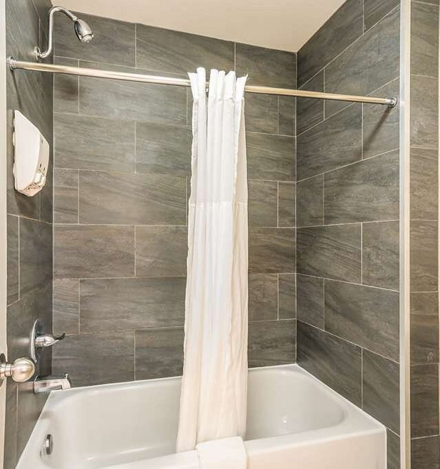 A small bathroom with a gray-tiled shower and bathtub, white shower curtain, towel rack with folded towels, and toilet.