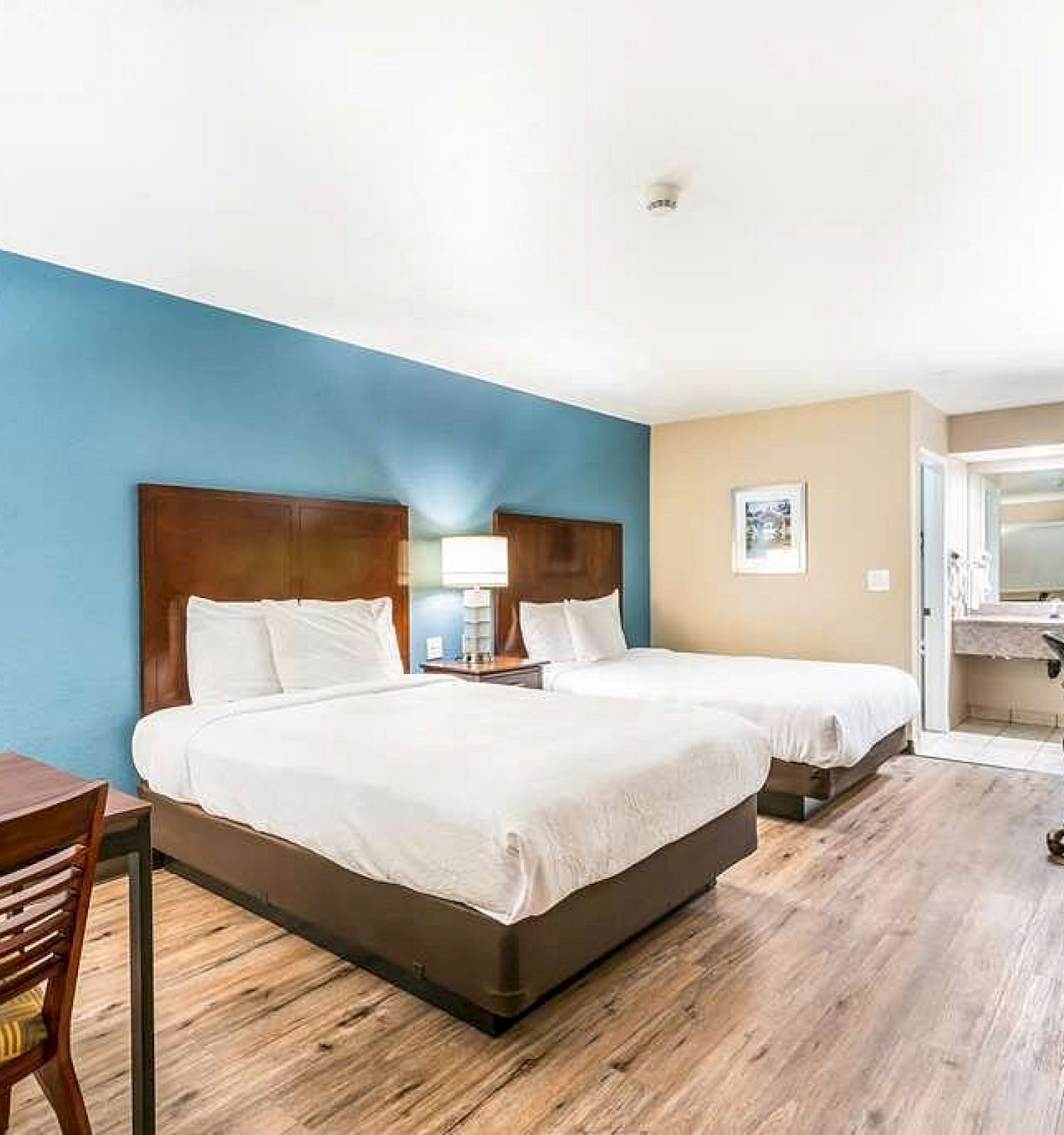 The image shows a hotel room with two double beds, a blue accent wall, a desk, a TV, and a bathroom in the background.