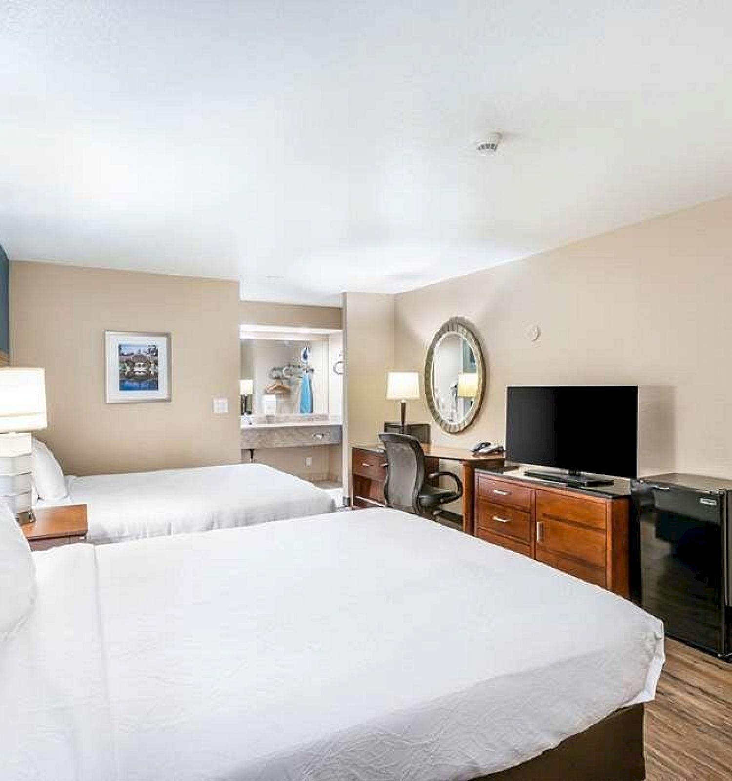 A hotel room with two beds, a TV, a refrigerator, a desk with a mirror, and a wardrobe in the background. The room has beige walls.