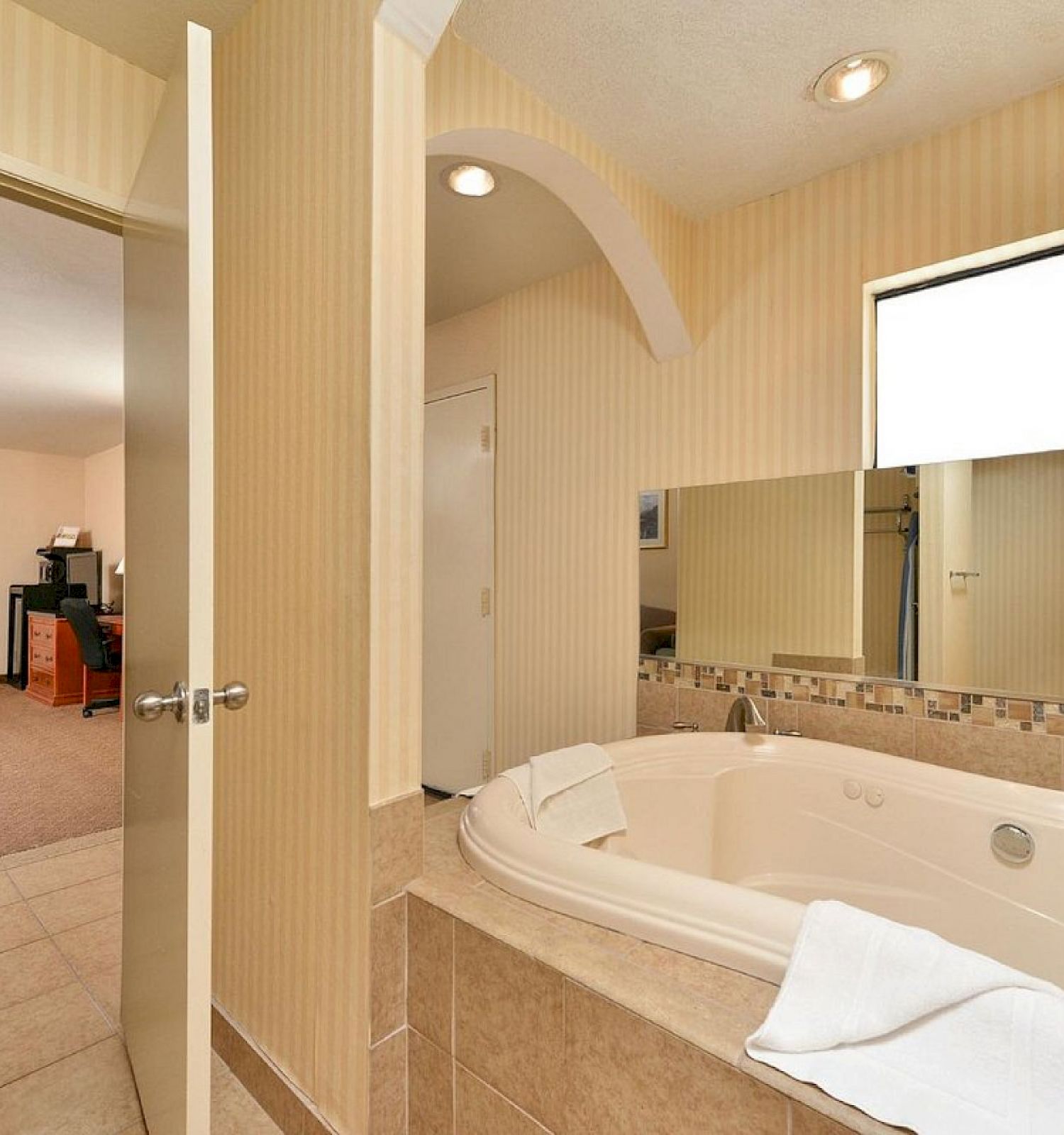 The image shows a hotel bathroom with a large bathtub and a view into the adjacent bedroom with beige tones and simple decor ending the sentence.