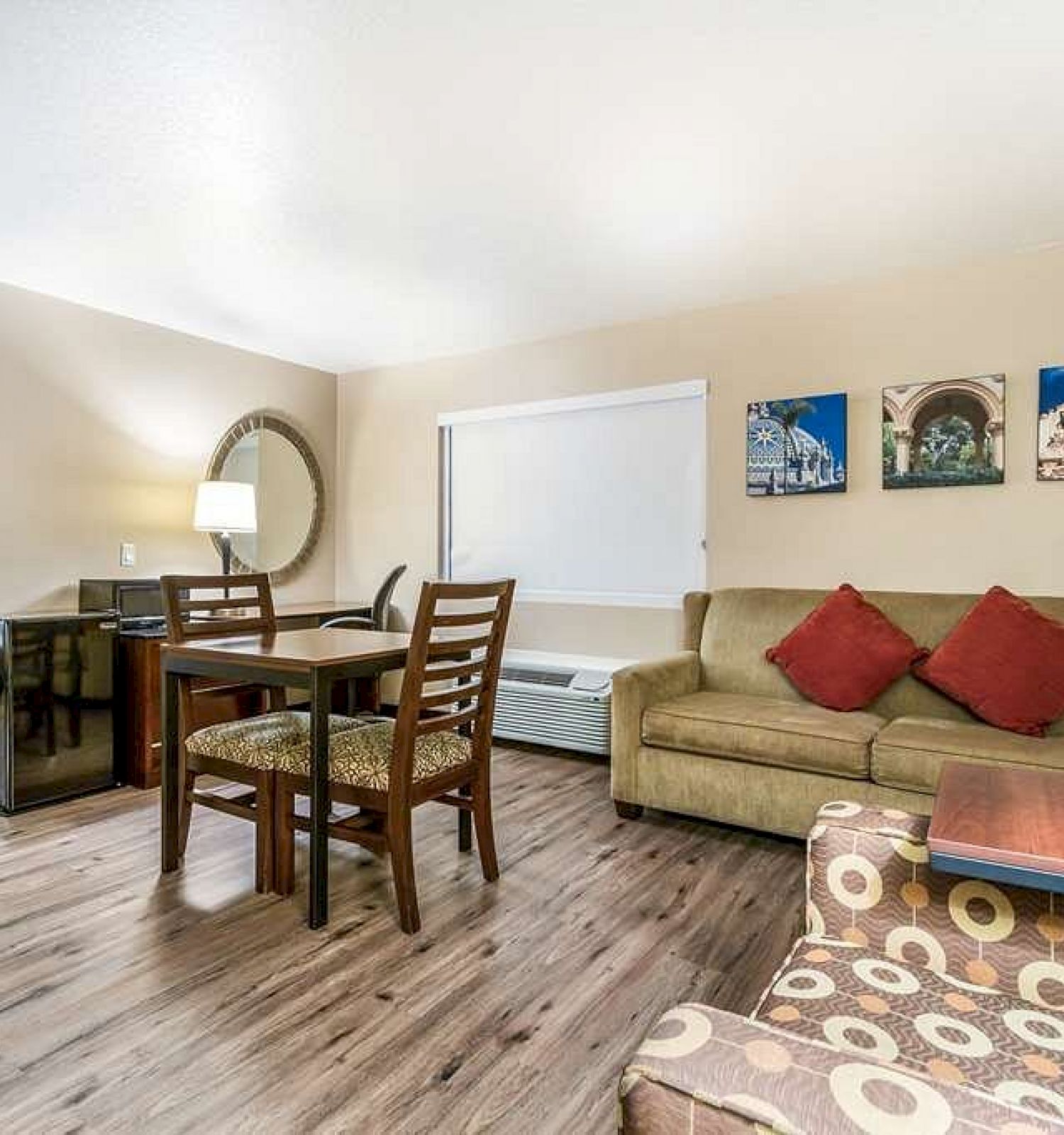 This image shows a living area with a couch, dining table, chairs, and an adjoining room with a bed. Wall art decorates the space.