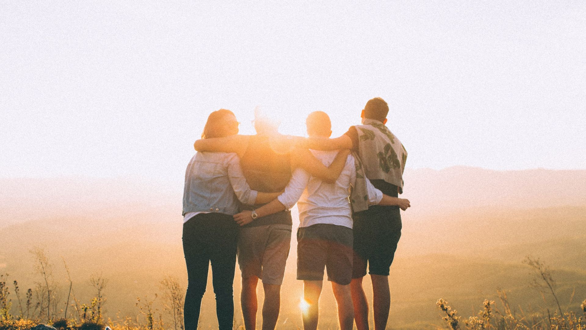 A group of four people stands on a hilltop, embracing each other while watching a beautiful sunset scenery in the background with bright sunlight.
