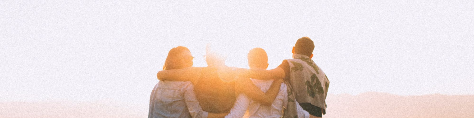 A group of four people stands on a hilltop, embracing each other while watching a beautiful sunset scenery in the background with bright sunlight.