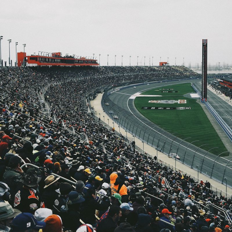 A large crowd fills the stands at a motor racing event, with the racetrack and infield visible under cloudy skies.