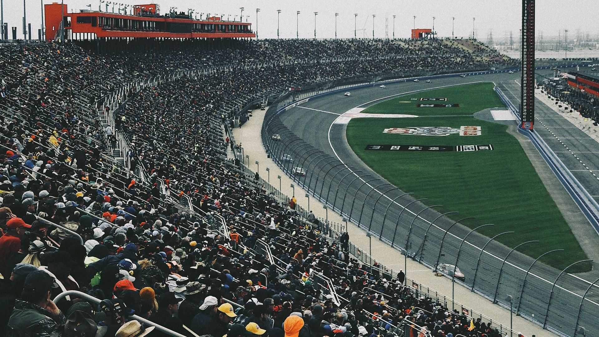 A large crowd fills the grandstands at a motorsport track, watching cars race around a curved section of the track, under an overcast sky.