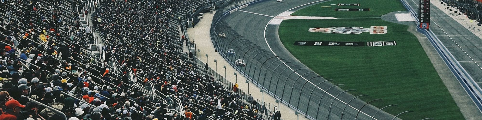 A large crowd fills the grandstands at a motorsport track, watching cars race around a curved section of the track, under an overcast sky.