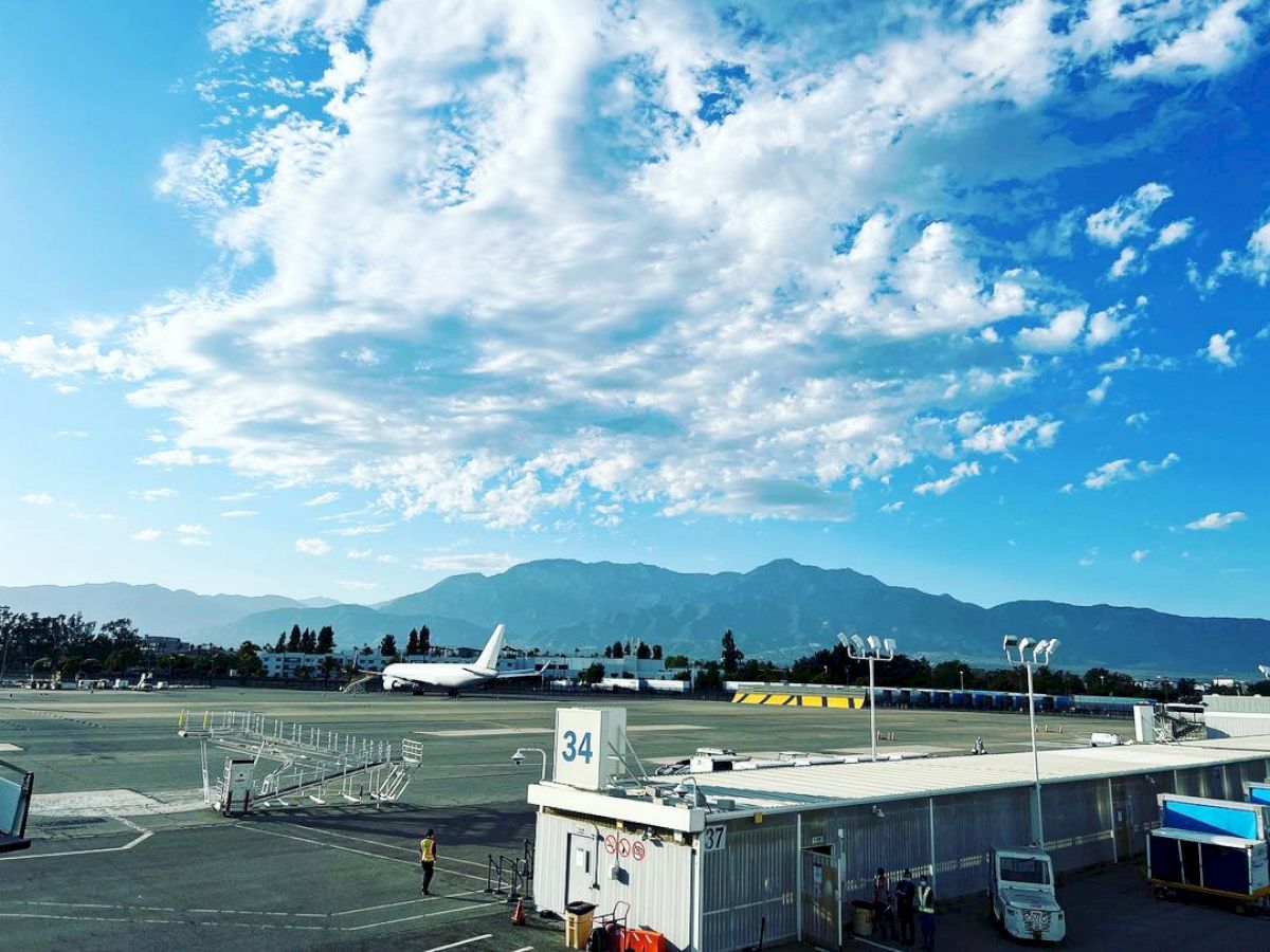 The image shows an airport runway with a parked airplane, a terminal building numbered 34, and a mountainous backdrop under a partly cloudy sky.