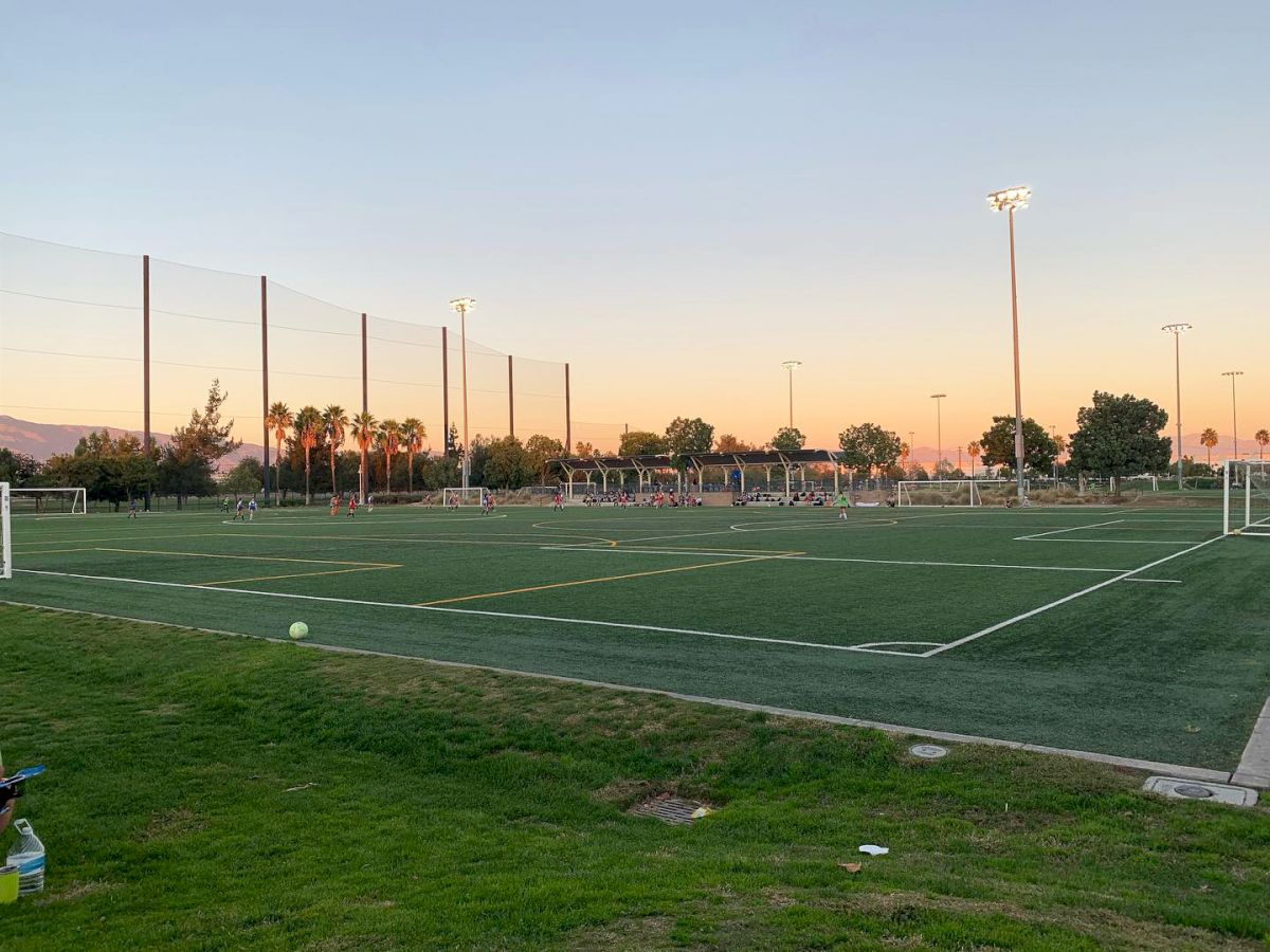 The image shows a soccer field during sunset with goalposts and surrounding lights. The area is empty, with a scenery of trees and a few people.