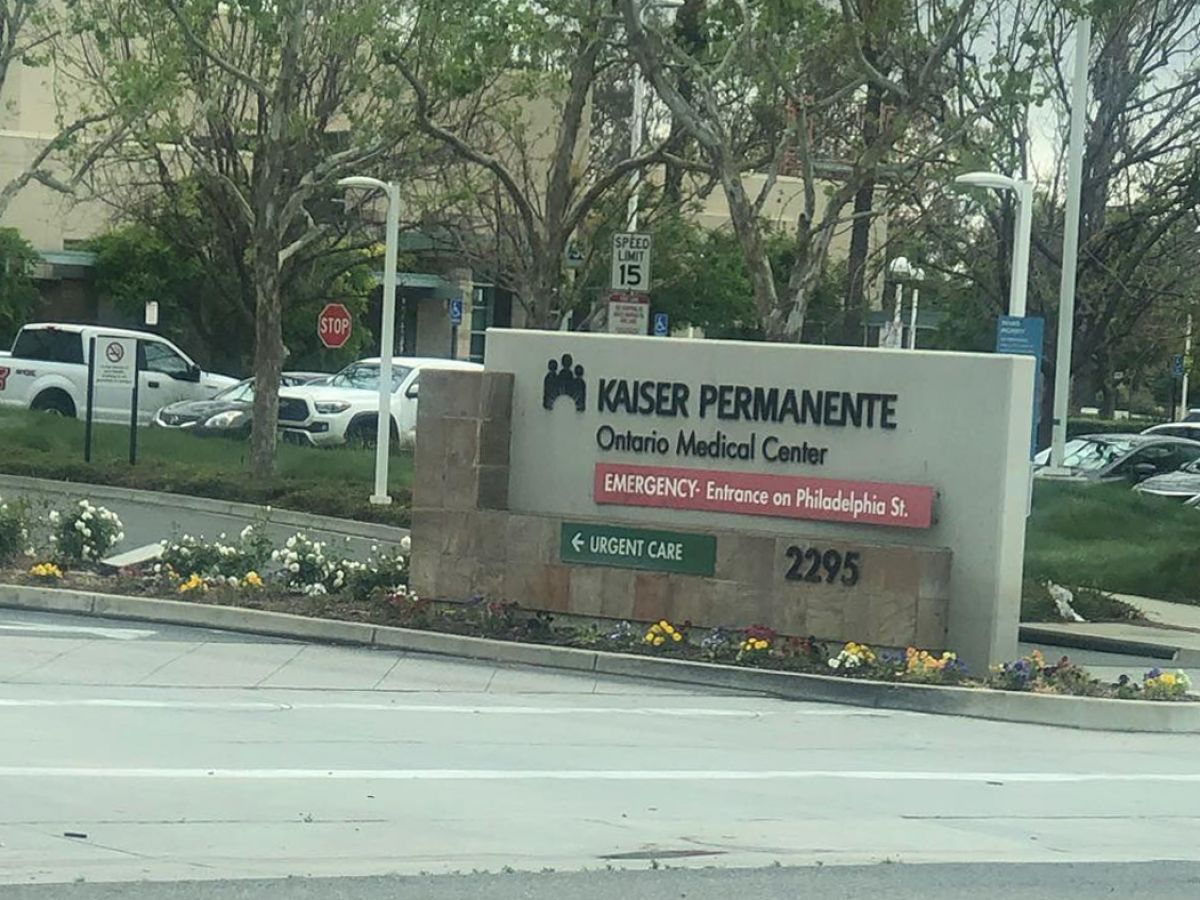 An outdoor scene with a sign for Kaiser Permanente medical center, indicating Emergency services and providing an address, with cars and greenery in the background.