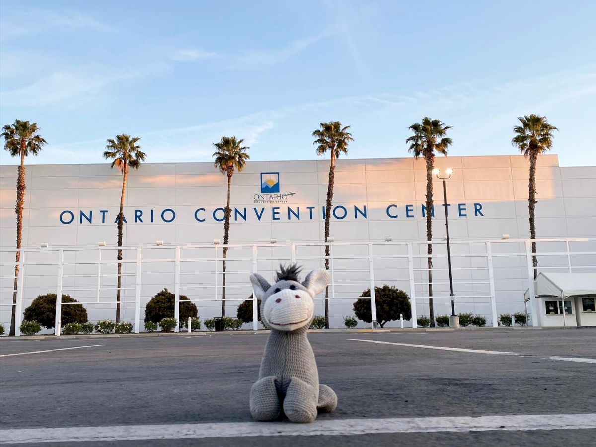 A stuffed donkey toy is placed in the foreground of an empty parking lot in front of the Ontario Convention Center building with palm trees around it.