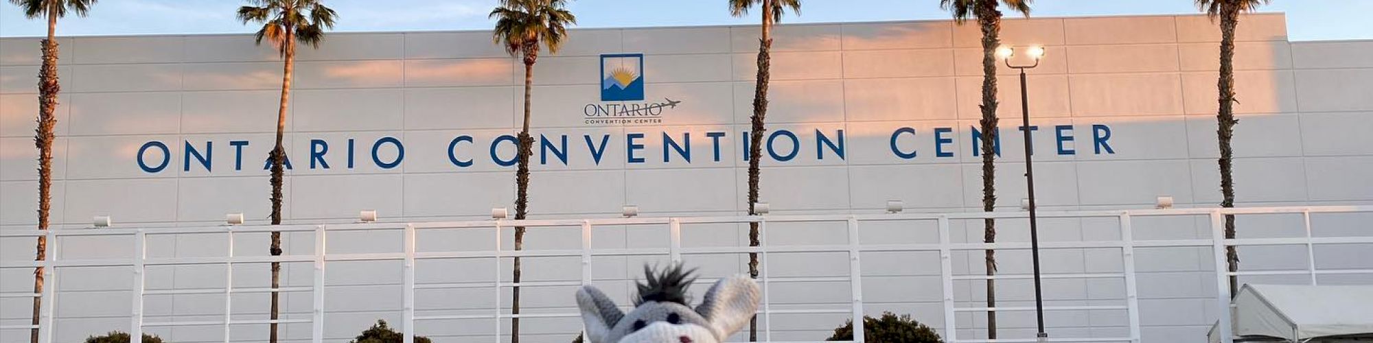 A stuffed donkey toy is placed on the ground in front of the Ontario Convention Center, with palm trees lining the background under a clear sky.