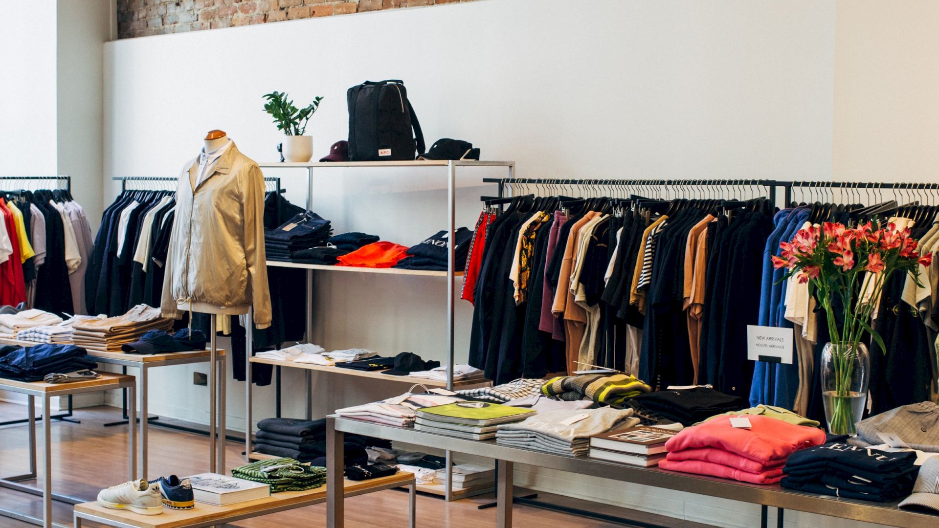 The image depicts a modern clothing store with racks of various garments, tables displaying folded clothes, and a brick accent wall.