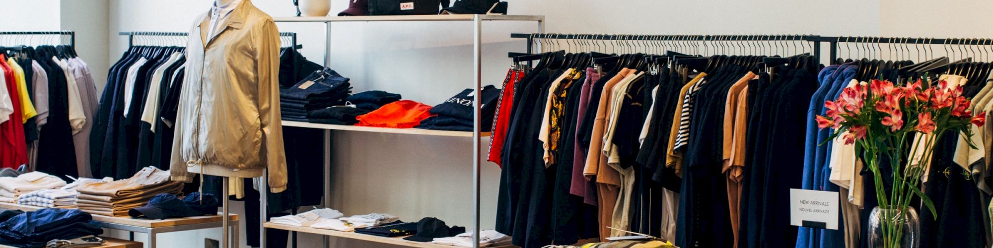 The image depicts a modern clothing store with racks of various garments, tables displaying folded clothes, and a brick accent wall.