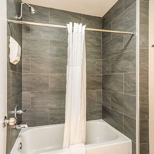 A bathroom with a gray-tiled bathtub and shower combo, a white shower curtain, and a towel rack holding folded white towels ends the image.