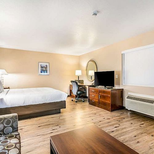 A hotel room with a bed, TV, desk with chair, armchair, and window air conditioning unit, featuring modern decor and a wooden floor.