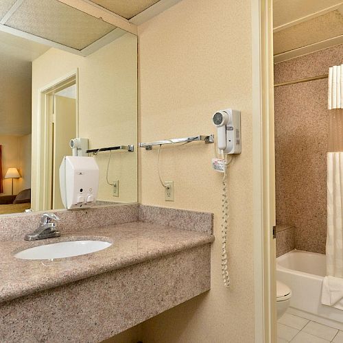 A bathroom with a sink, mirror, and hairdryer, next to a bathtub with a curtain. Nearby, an open doorway leads to a room with a window and lamp.