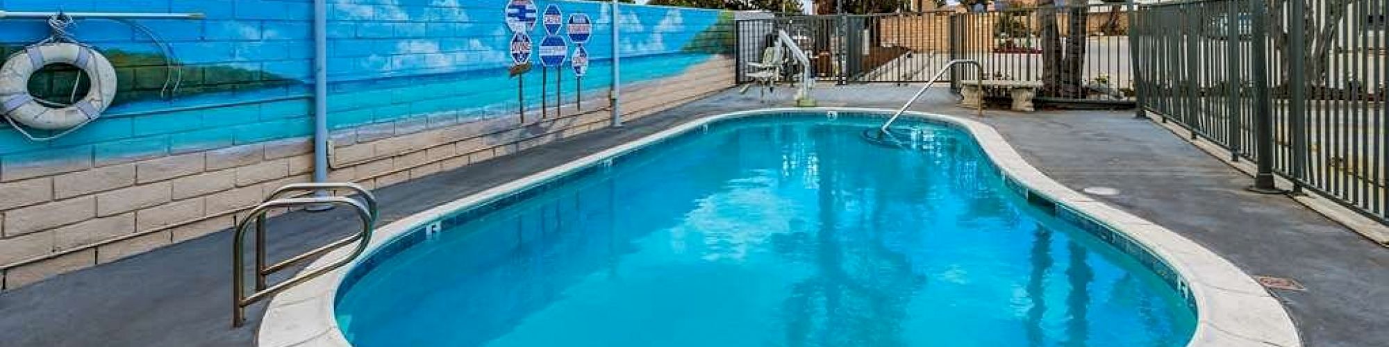 An outdoor swimming pool enclosed by a fence, with clear water, a painted mural on the wall, and safety equipment visible in the background.