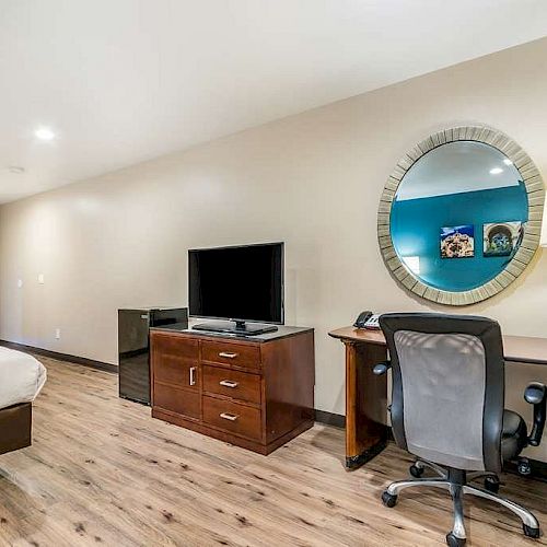 The image shows a modern hotel room with a bed, a TV on a wooden dresser, a desk with a chair, a mirror, a lamp, and wooden flooring, ending the sentence.