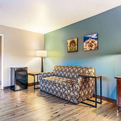 A modern living room features a patterned sofa, wooden floors, two lamps, a side table, and wall art, with a view into a bathroom.