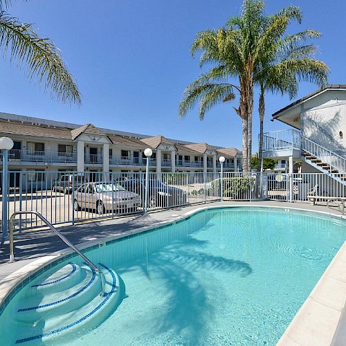 A small, gated swimming pool is in front of a two-story motel with parked cars and palm trees under a clear blue sky.