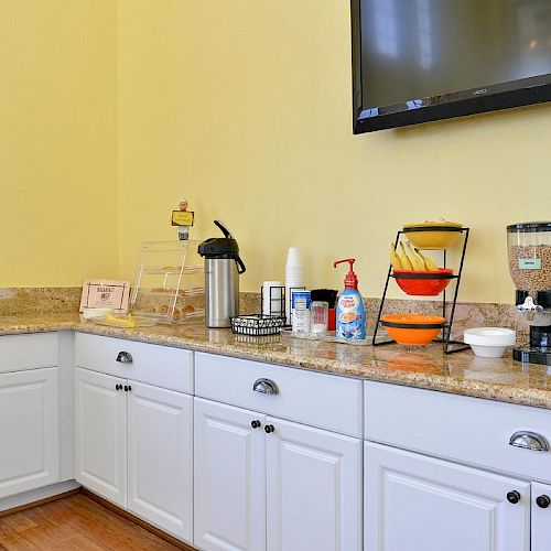 The image shows a kitchenette area with white cabinets, a microwave, cereal dispensers, coffee station, fruit, a TV on the wall, and countertop items.