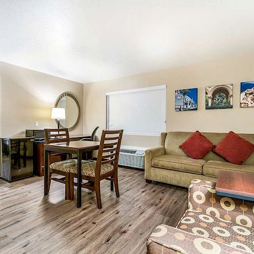 A cozy living area with a sofa, armchair, dining table, and chairs. Adjacent bedroom with a bed, lamp, and artworks on the wall above the sofa.