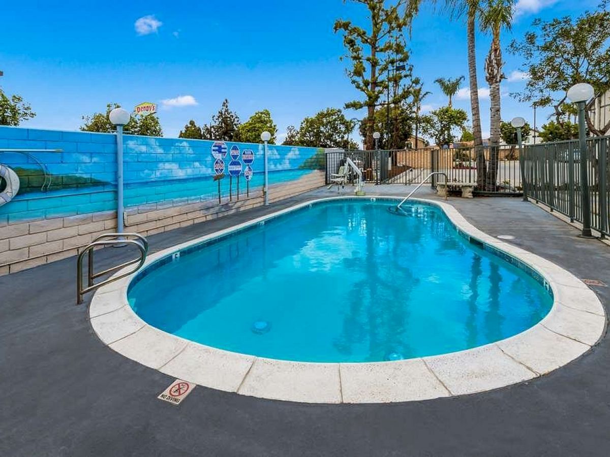 An outdoor swimming pool surrounded by a fence, with safety signs and a mural on the wall, set in a sunny location with trees and a clear blue sky.