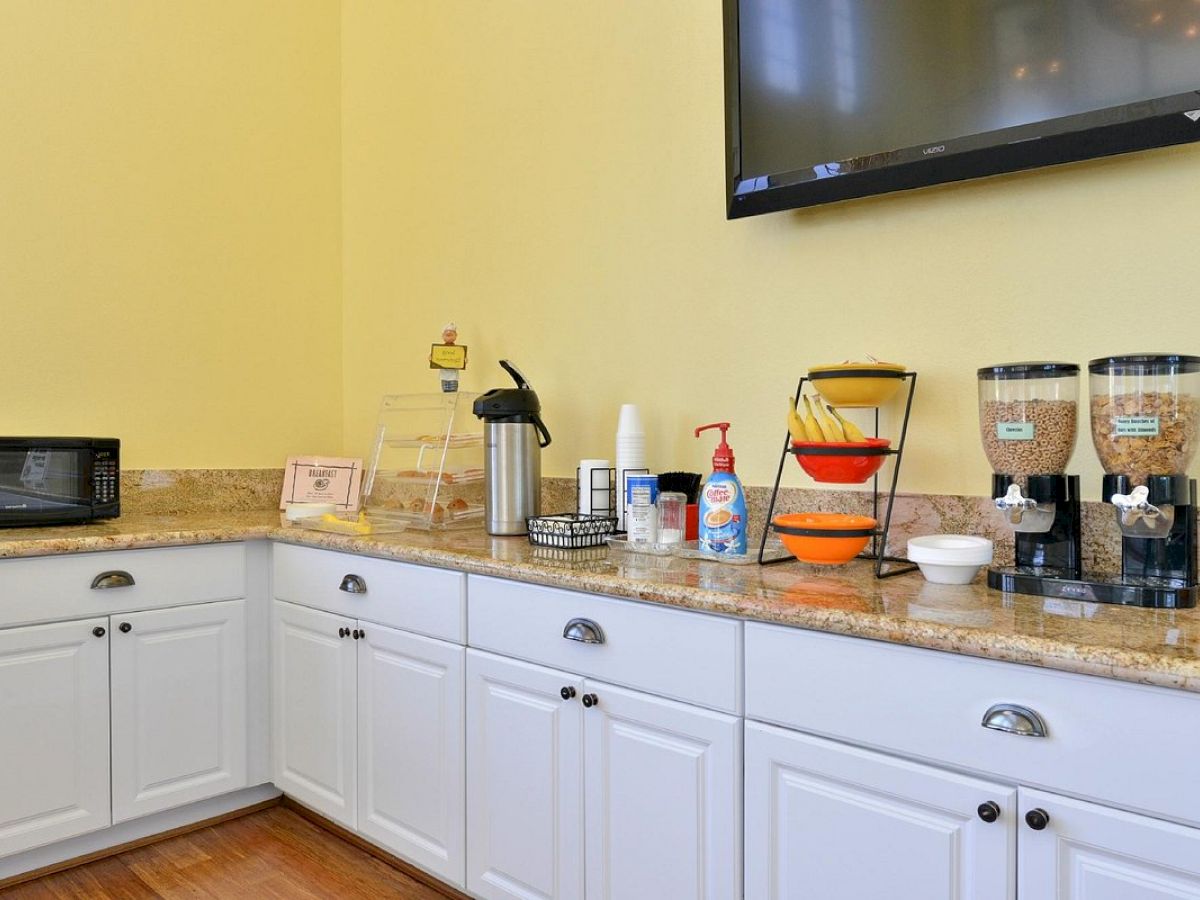 A kitchen area with a microwave, beverage dispenser, cereal dispensers, fruit, utensils, and a TV mounted on the wall ends the sentence.