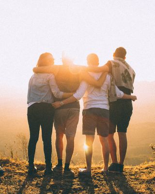 Four people stand together on a hilltop at sunset, with their arms around each other, facing the horizon and enjoying the scenic view.