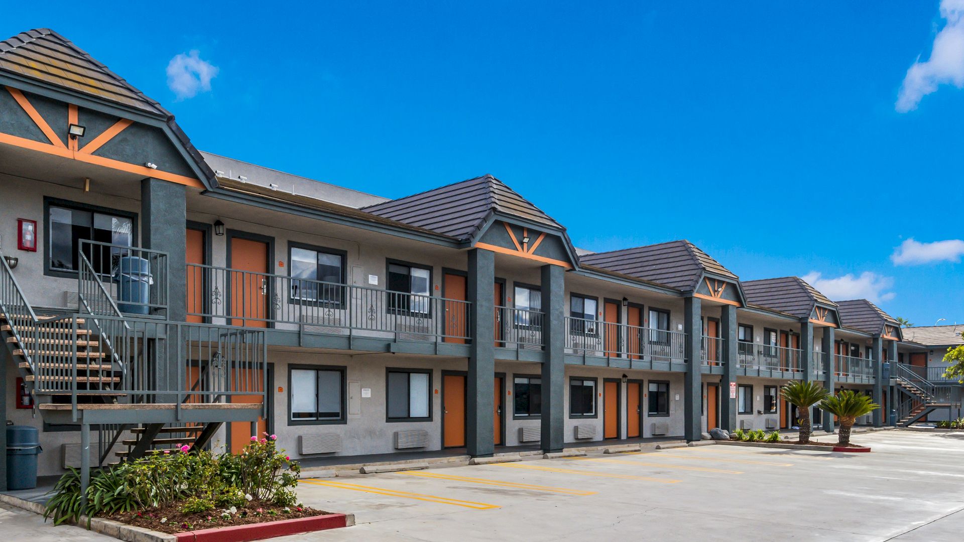A two-story motel building under a clear blue sky, with parking spaces and small garden beds visible in the foreground.