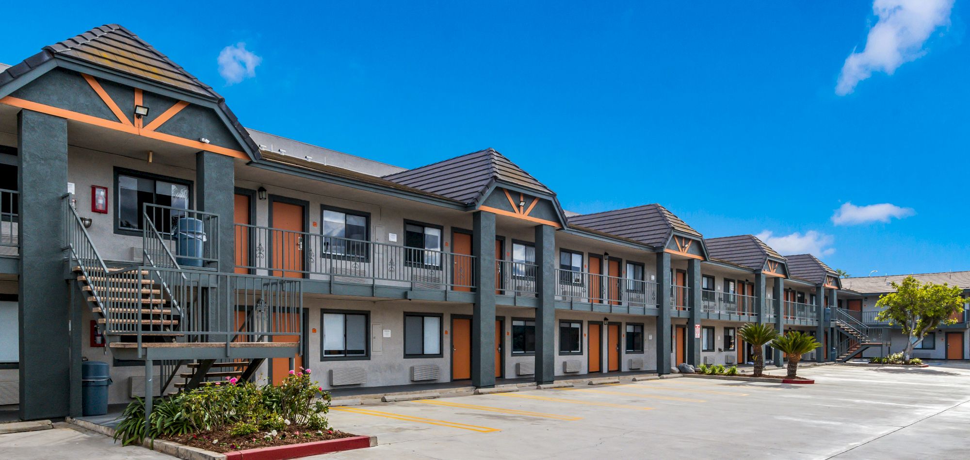 A two-story motel building under a clear blue sky, with parking spaces and small garden beds visible in the foreground.