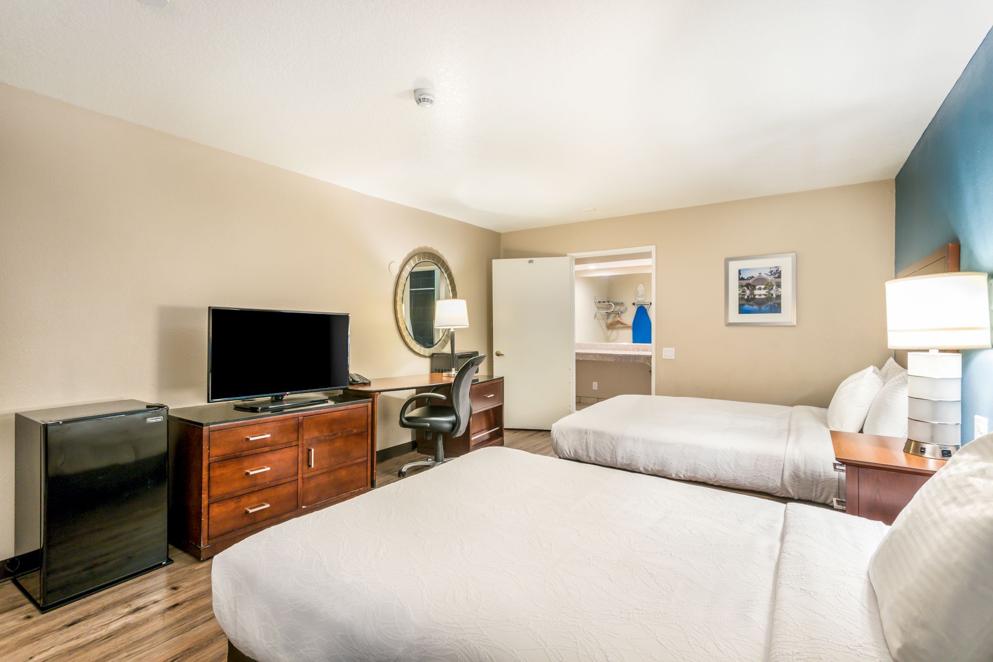 A hotel room with two beds, a TV on a dresser, a mini-fridge, a desk with a chair, and a mirror on the wall. The room has wooden flooring.