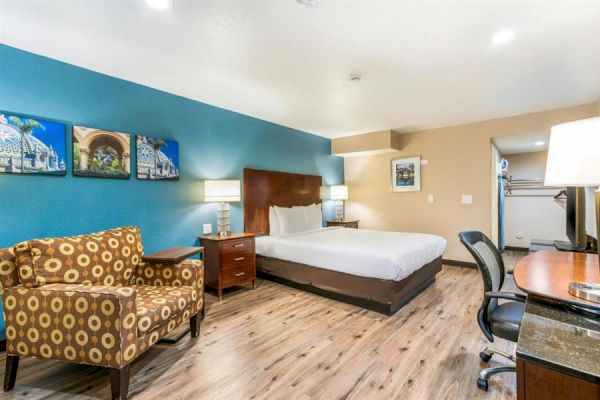 A hotel room featuring a large bed, colorful armchair, desk with chair, artwork on the wall, lamps, and a wooden floor.