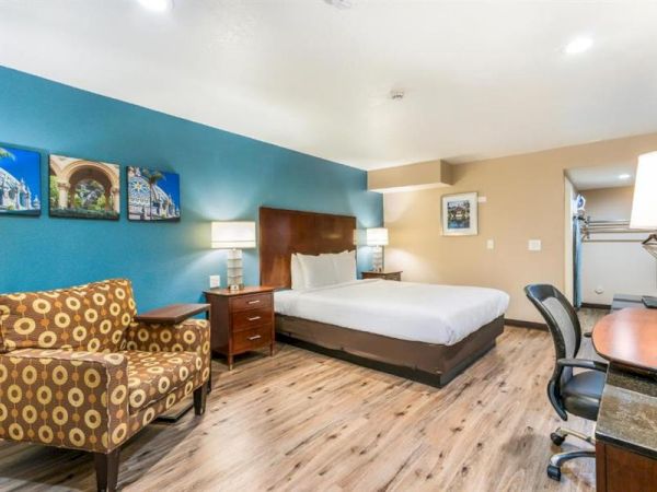 A hotel room featuring a large bed, colorful armchair, desk with chair, artwork on the wall, lamps, and a wooden floor.