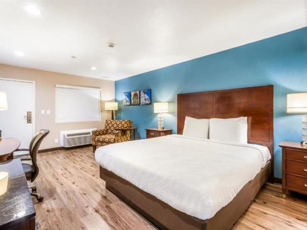 A hotel room with a large bed, two nightstands with lamps, a desk and chair, an armchair, artwork on the wall, and wood flooring, seen.