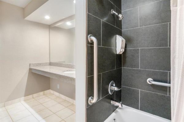 The image shows a modern bathroom with a tiled shower area, grab bars, and a soap dispenser. Adjacent to it is a countertop sink with a mirror.