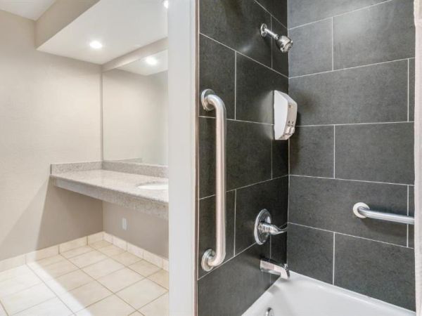 The image shows a modern bathroom with a tiled shower area, grab bars, and a soap dispenser. Adjacent to it is a countertop sink with a mirror.