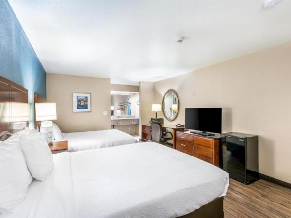 The image shows a hotel room with two beds, a TV on a dresser, a small fridge, a desk with a chair, and a mirror above the desk.