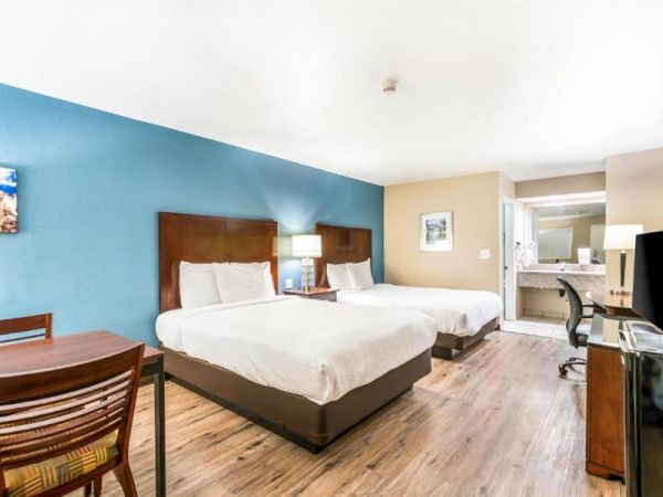 The image shows a hotel room with two beds, a desk with chair, a TV, a nightstand with a lamp, and a bathroom area in the background.