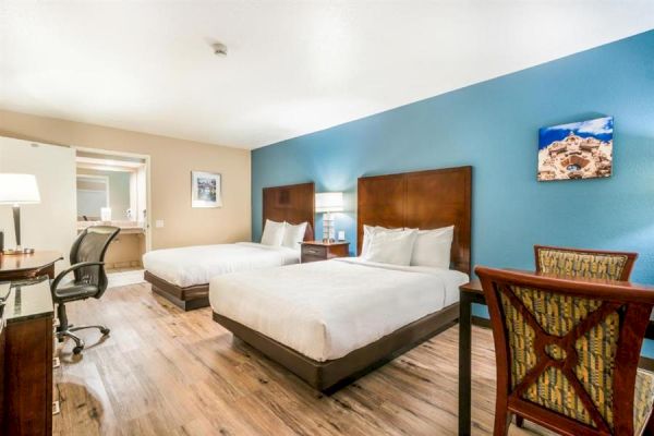 The image shows a hotel room with two double beds, wooden floors, a workspace with a chair, a small table, and wall art above each bed.