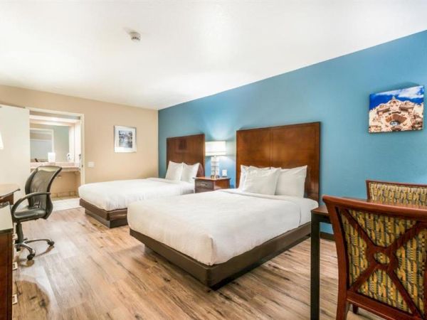 The image shows a hotel room with two double beds, a blue accent wall, a desk with a chair, a small round table with chairs, and a wall picture.