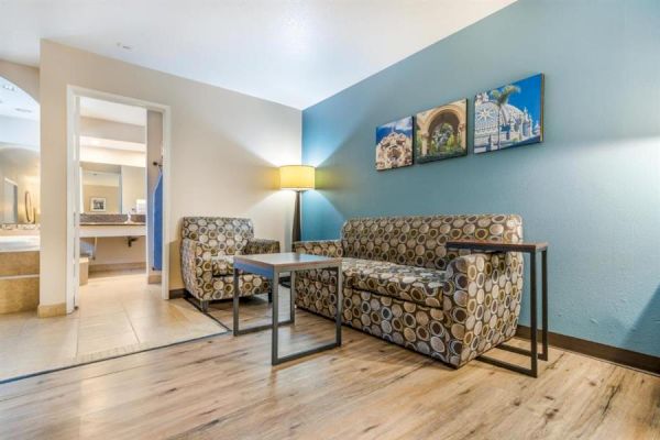 A modern hotel room sitting area with patterned couches, a lamp, artwork on the wall, and an open bathroom door in the background.