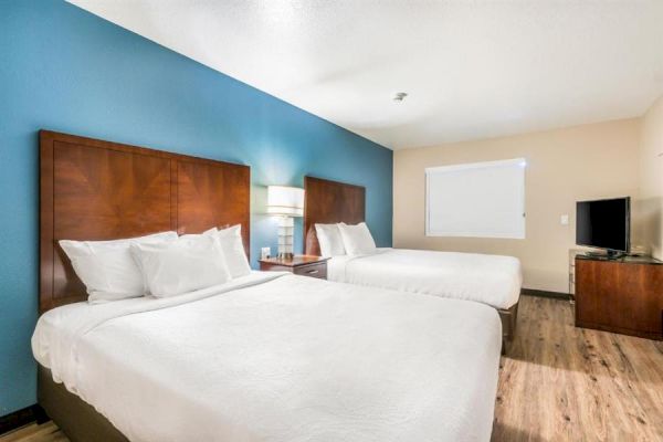 The image shows a hotel room with two beds, a nightstand with a lamp, a TV on a wooden dresser, and a window with a closed blind.