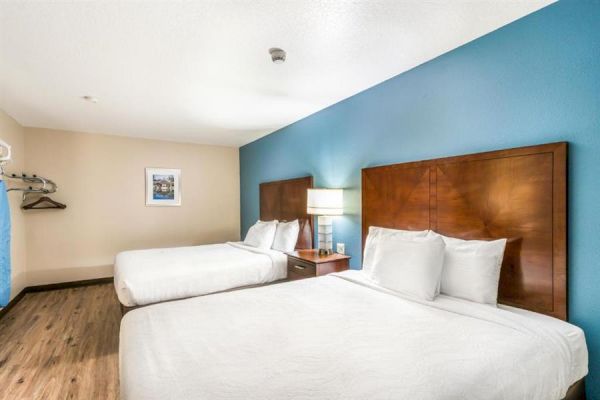 The image shows a hotel room with two double beds, a nightstand with a lamp, a coat rack, and a framed picture on the wall.