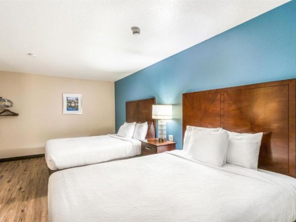 The image shows a hotel room with two double beds, a nightstand with a lamp, a coat rack, and a framed picture on the wall.